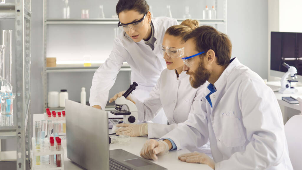 Three lab technicians in white coats and safety glasses reviewing data on a laptop next to a microscope in a laboratory setting