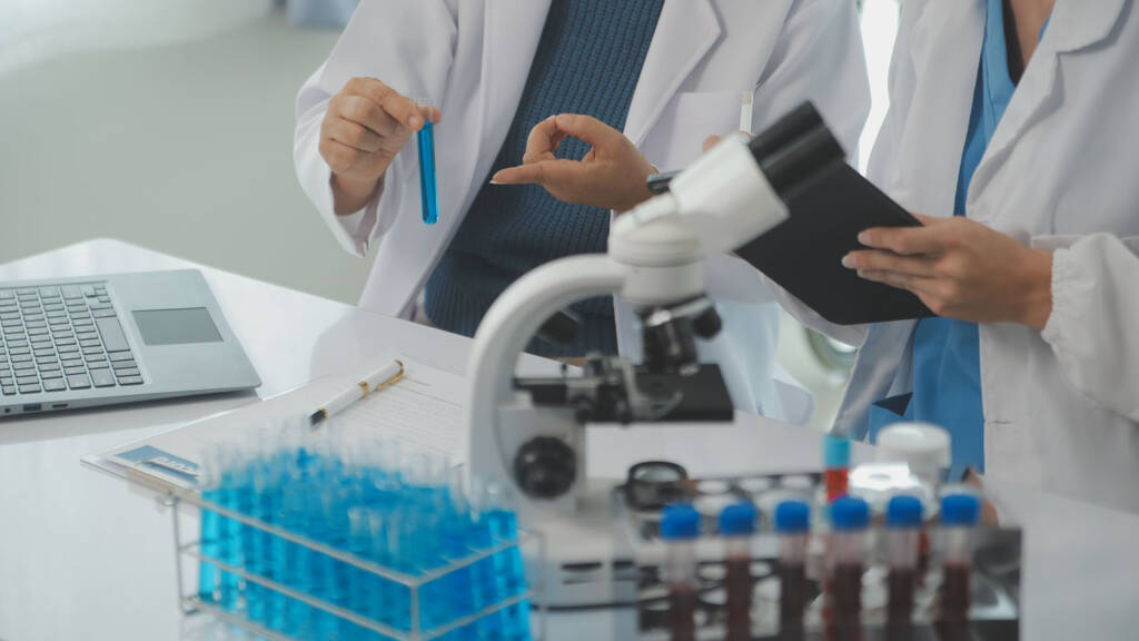 Two laboratory professionals in white coats reviewing a sample vial next to a microscope, laptop, and rack of reagent tubes