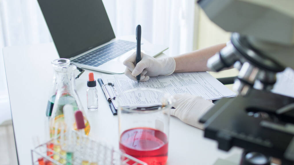 Laboratory professional in white gloves manually recording test results on paper forms surrounded by reagents and a microscope, highlighting the need for automated LIMS data management