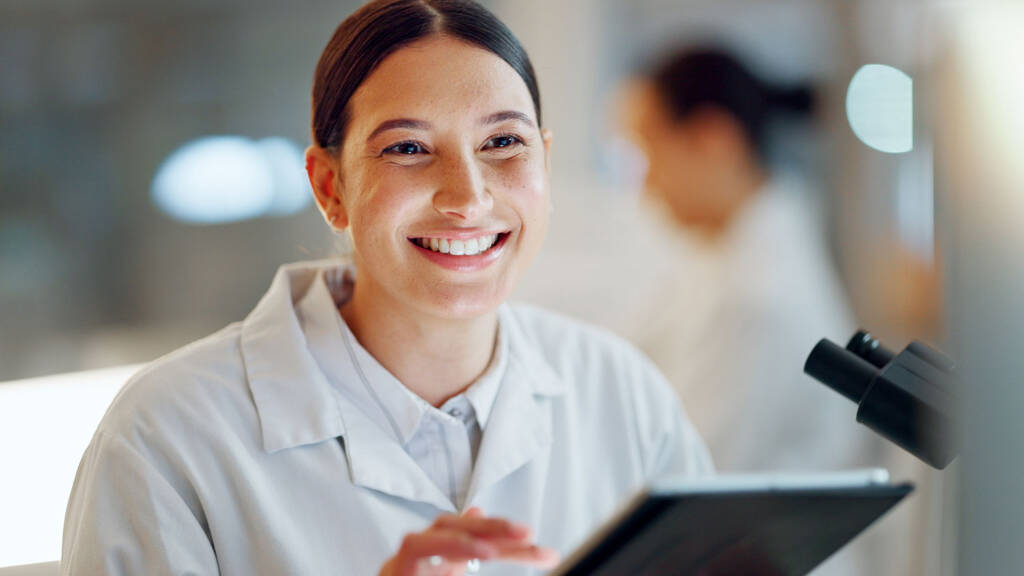 Smiling female lab professional in a white coat holding a tablet in a modern laboratory, representing efficient data access enabled by LIMS software