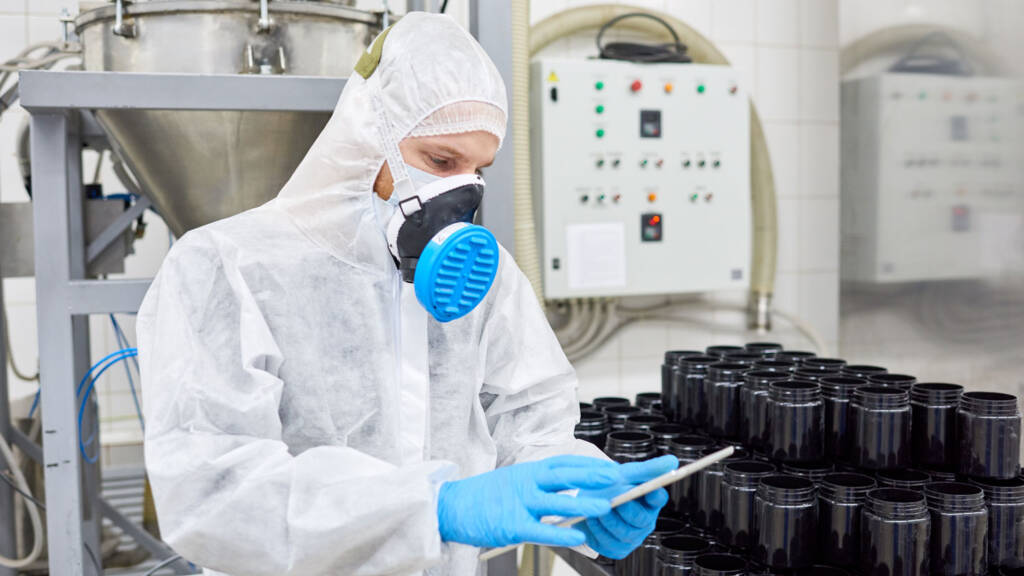 Laboratory worker in protective equipment examining a sample near stored containers in an industrial testing facility.
